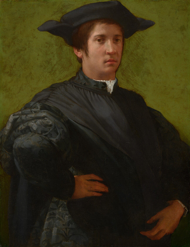This painting shows a young man against an olive-green background. The man is standing, his body partially turned to the right, with one hand on his hip and the other near his waist. He is looking at us with a neutral expression. The man has light skin and short brown hair under a black hat with pointed ends on either side. He wears a dark outfit with a high white collar and puffed black sleeves. A thin gold ring is visible on his right hand. The green background has visible brushstrokes and some slight variation in color.