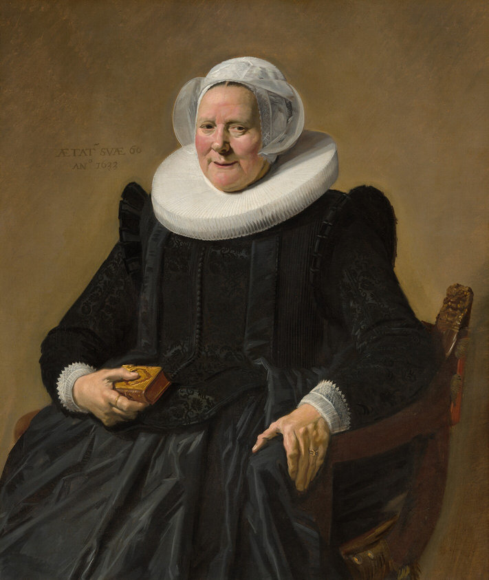 This vertical portrait painting shows a pale-skinned older woman wearing a full, long-sleeved black dress with a wide white ruff at her neck and white lace cuffs at her wrists. She sits in a curving, low-backed wood chair against a pale brown background. Her body and the chair are angled slightly to our left, and she looks directly at us. She has small eyes, pink, rounded cheeks, and a wide chin. Her lips are parted in a slight smile, and her hair is covered by a starched, sheer white cap that flares at the sides. The ruff around her neck is gathered in accordion-like, narrow figure-eight folds, and it extends flat and stiff, nearly to her shoulders. Close inspection reveals that the dress is woven with a black-on-black brocade pattern and has a line of small black buttons down the front. She holds a small brown leather book tooled with gold ornament in her right hand, on our left, and her other hand rests on the arm of the chair. She wears a gold ring on each hand. An inscription is painted to our left of her head: “AETAT SVAE 60 ANo 1633.”
