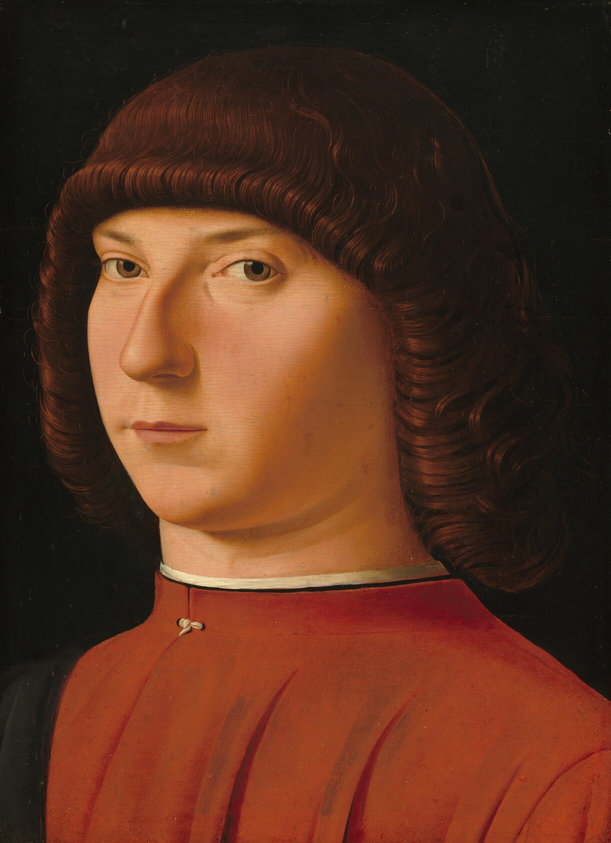 Shown from the chest up, a clean shaven, young man with pale, rosy skin and curly, mahogany-red hair, looks at or toward us against a black background in this vertical portrait painting. His body is angled to our left, and he turns his face slightly to look our way with sienna-brown eyes under curved eyebrows. He has a long, prominent nose and small thin pink lips. His shoulder length hair curls tightly around his face and covers his ears. He wears a tomato-red shirt with a high collar trimmed with white.