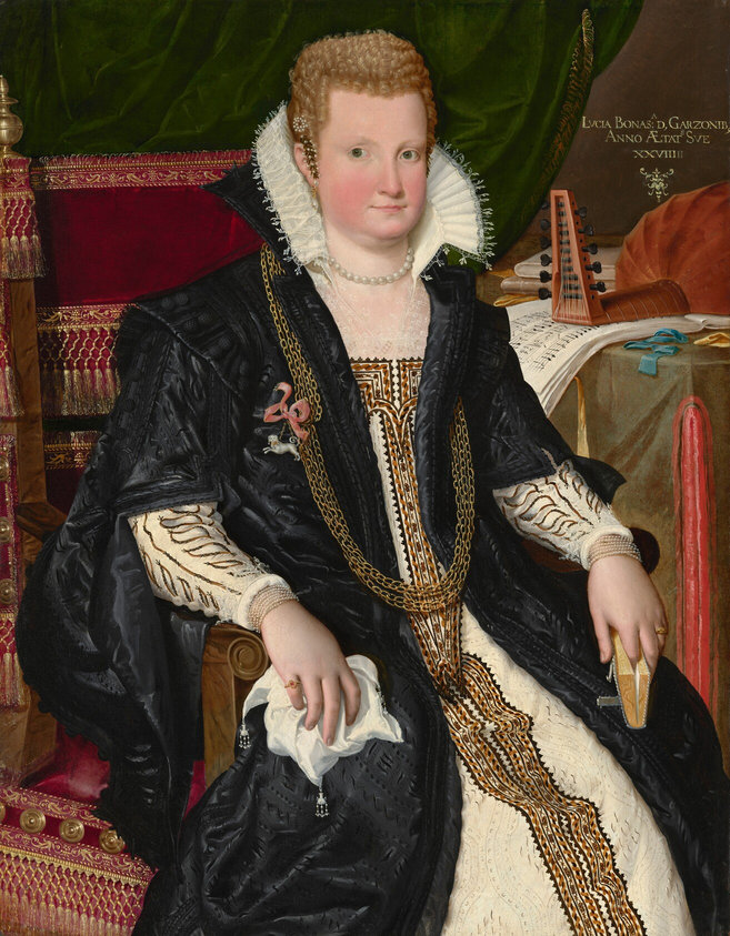 This painting is a portrait of a seated woman, her body turned slightly to the right and her head looking towards us. She has a fair complexion, with round, rosy cheeks, pale eyebrows, large green eyes, and thin pink lips in a neutral expression. Her light blonde hair is styled in short curls close to her head and adorned with pearls and a clip or barrette in the shape of small leaves. She is dressed in a cream-colored dress with intricate embroidery in shades of brown, gold, and black. On top of the dress is a long black garment that flows over her shoulders and upper arms and has a high collar, decorative buttons, and more embroidery. Her gown is paired with a ruffled white collar and lace at her neckline and wrists. Long gold chains drape across her chest, and she wears a pearl necklace around her neck and two small golden rings on her fingers. In her left hand, our right, she holds a small book, while a lace handkerchief is in her right hand, our left. She wears pearl earrings, strands of pearl bracelets, and a decorative brooch in the shape of a white dog with a pink bow. The seat she sits on is wooden with dark red cushions decorated with golden tassels and embroidery, behind her on the left is a draped green cloth, and on the right, a table is covered with music sheets, a stringed instrument, books, and small blue and yellow ribbons. The wall behind the table is brown, and there is some gold lettering in the top right corner.